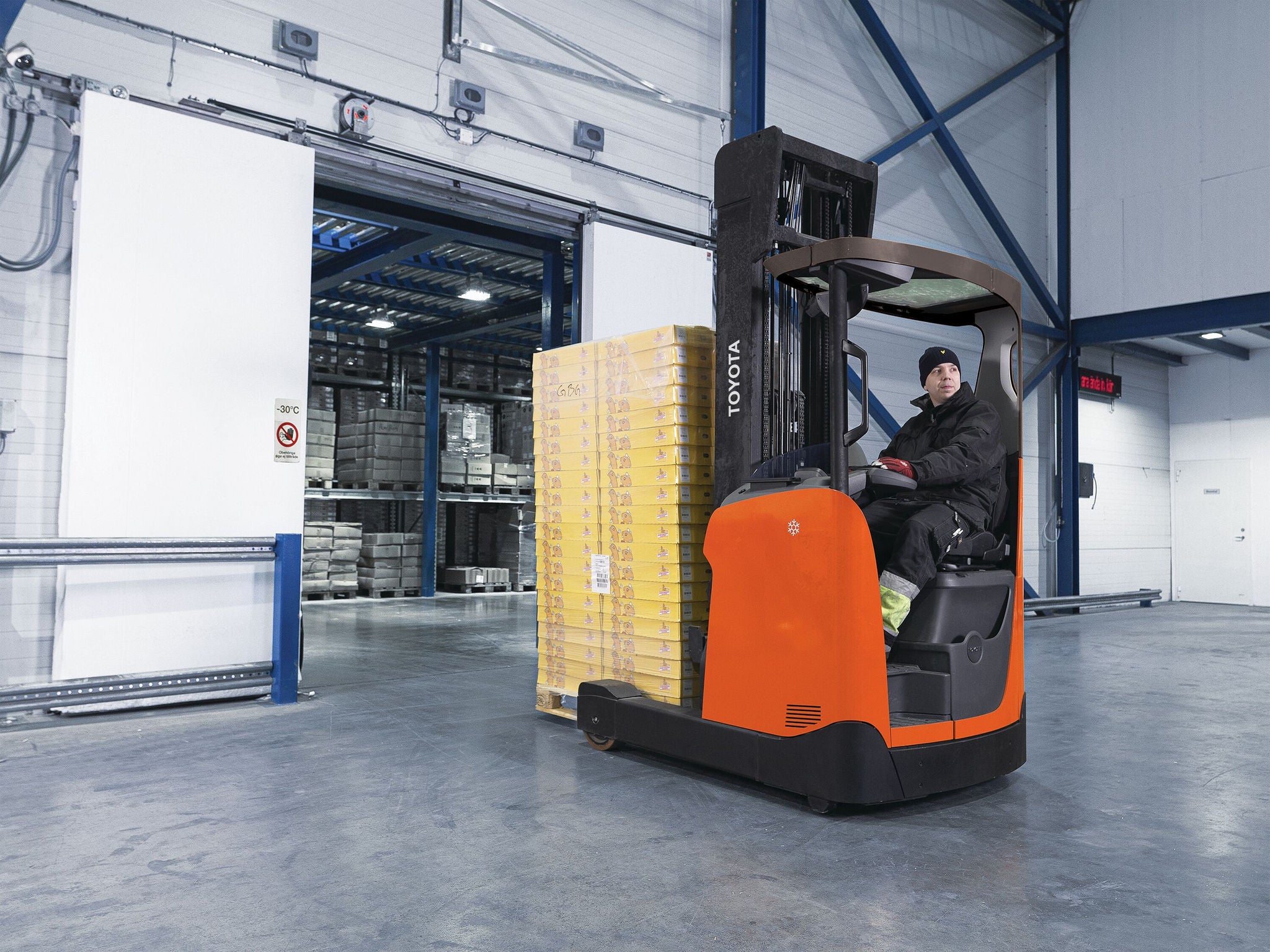 Man driving Toyota Moving Mast reach truck carrying tall stack of pallets in cold storage warehouse 